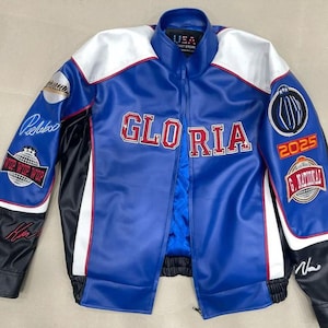 Puede incluir: Chaqueta azul y blanca de cuero sintético con la palabra "GLORIA" en letras rojas. La chaqueta tiene cremallera frontal, mangas negras y parches con texto y gráficos. La chaqueta tiene cuello alzado y ribetes rojos.
