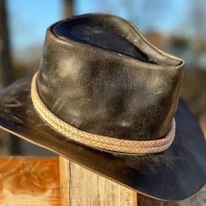 May include: A black leather cowboy hat with a braided band. The hat has a wide brim and a slightly distressed appearance. The hat is resting on a wooden fence.