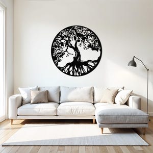 May include: A black metal wall art sculpture of a tree of life with intricate branches and roots. The sculpture is circular and is mounted on a white wall above a white couch.