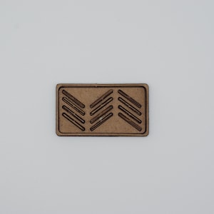 May include: A rectangular wooden plaque with a series of diagonal lines carved into its surface. The lines are arranged in a chevron pattern, with three sets of lines pointing downwards. The plaque is a light brown color.
