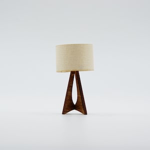 Könnte beinhalten: Eine Miniatur-Tischlampe mit beigem Stoffschirm und dunkelbraunem Holzfuß. Der Fuß hat ein einzigartiges dreieckiges Design. Die Lampe ist ein dekoratives Accessoire, perfekt für ein Puppenhaus oder eine Miniaturumgebung.