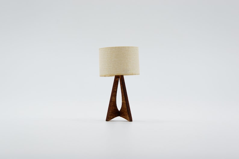 May include: A miniature table lamp featuring a cylindrical, off-white fabric lampshade resting on a dark brown, triangular wooden base with a hollow center. The lamp has a smooth, polished finish and stands upright on a plain, neutral surface.