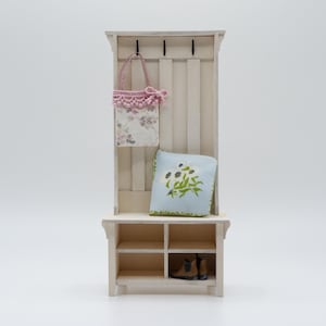 Könnte beinhalten: Ein Miniatur-Garderobenständer aus weißem Holz mit einem Regal, Haken und einem kleinen Stauraum. Eine Tasche mit floralem Muster und rosa Spitzenbesatz hängt an einem Haken. Ein hellblaues Kissen mit grünem Blumendesign liegt auf dem Regal.