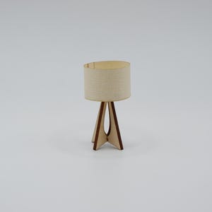 May include: A miniature table lamp featuring a cylindrical beige fabric lampshade and a tripod base constructed from thin, dark-edged light brown wood. The lamp has a simple, open-frame design with a central vertical support and three legs.