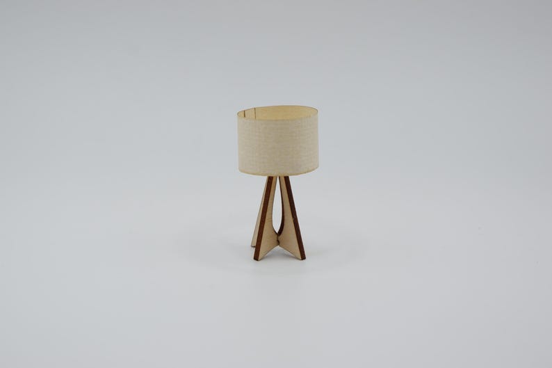 May include: A miniature table lamp featuring a cylindrical beige fabric lampshade and a tripod base constructed from thin, dark-edged light brown wood. The lamp has a simple, open-frame design with a central vertical support and three legs.