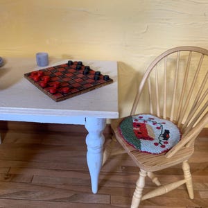 May include: Miniature scene featuring a checkerboard with red and black checkers on a white table with blue legs. A wooden chair with a round cushion featuring a bird design sits nearby. The scene is set against a yellow wall.