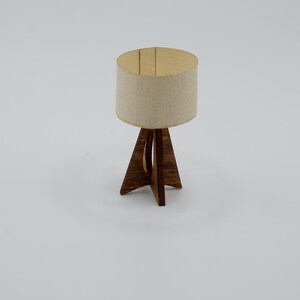 May include: A miniature table lamp featuring a cylindrical off-white fabric lampshade and a dark brown wooden base with three intersecting legs that form a tripod structure. The lamp has a simple, clean design with visible wood grain texture.