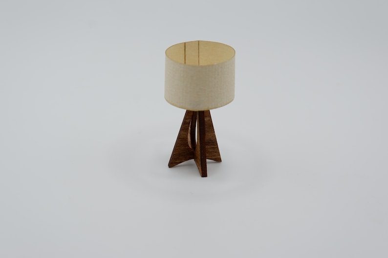 May include: A miniature table lamp featuring a cylindrical off-white fabric lampshade and a dark brown wooden base with three intersecting legs that form a tripod structure. The lamp has a simple, clean design with visible wood grain texture.