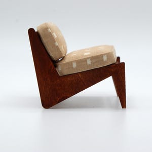 May include: A miniature lounge chair featuring a dark brown wooden frame with an angular profile. The seat and backrest are upholstered in a light beige fabric with a repeating white square pattern, providing a contrast to the dark wood structure.