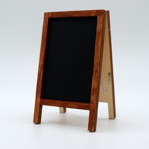 May include: A small A-frame chalkboard sign featuring a dark brown wooden frame and a black writing surface. The sign is constructed with two hinged panels connected by a thin silver metal chain on the side to maintain its standing position.