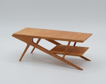May include: A miniature wooden coffee table featuring a rectangular top and a lower shelf. The table has angled, tapered legs that extend outward from the frame, creating a distinct geometric profile with a light brown wood grain finish.