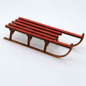 May include: A miniature wooden sled with a red seat and brown runners. The sled has a classic design with curved runners and a slatted seat. Perfect for holiday decor or a winter-themed craft project.