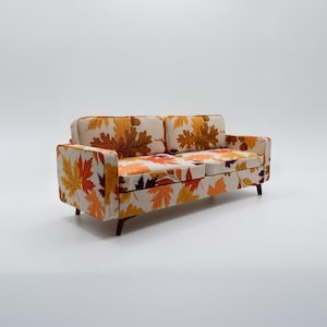 Könnte beinhalten: Ein Zweisitzer-Sofa mit cremefarbenem Hintergrund und einem Muster aus Herbstblättern in Orange-, Gelb- und Brauntönen. Das Sofa hat eine rechteckige Form mit niedriger Rückenlehne und Armlehnen und steht auf vier Holzbeinen.