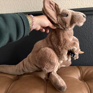 May include: A plush kangaroo toy with soft brown fur, large upright ears, and a long tail. The kangaroo features a pouch on its front containing a small joey, which is also made of brown plush material and has visible black plastic eyes.