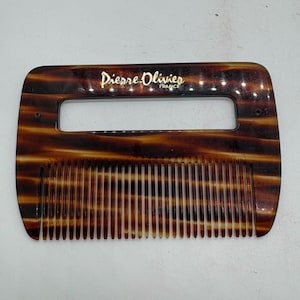 Könnte beinhalten: Ein Vintage-Kamm von Pierre Olivier mit Schildpattmuster. Der Kamm hat einen rechteckigen Griff mit Aussparung und feinen Zähnen. Der Markenname "Pierre Olivier FRANCE" ist in goldfarbenen Buchstaben auf dem Griff aufgedruckt.