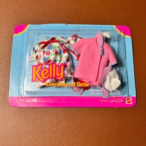 Op de afbeelding: Een verpakte poppenkledingset met een jurk met bloemenprint en rode strik, een roze badjas en witte pantoffels. De verpakking is blauw en roze, met de tekst "Kelly Baby Sister of Barbie".