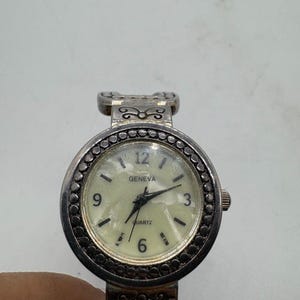May include: A silver-toned Geneva Quartz watch with a round face. The watch has a cream-colored dial with black numerals and hands. The bezel is decorated with small, dark-colored beads. The watch band is also silver-toned with decorative elements.