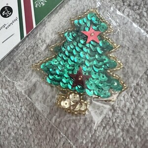 Peut inclure: Ornement de sapin de Noël fait de paillettes vertes avec une bordure dorée perlée. L'arbre est surmonté d'une étoile rouge et est emballé dans un sac en plastique transparent. L'étiquette indique "wings... everyday!"