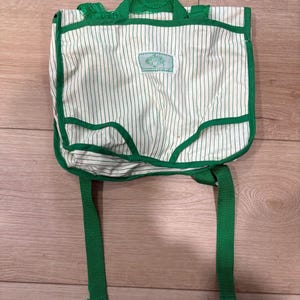May include: A white backpack with thin vertical green stripes, featuring green fabric trim, a top handle, and two adjustable shoulder straps. A small rectangular white label with green text and a graphic is centered on the front flap of the bag.
