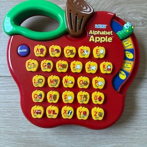 Peut inclure: Un jouet éducatif électronique VTech Alphabet Apple en forme de pomme rouge avec une poignée verte et une tige marron. Il comporte des boutons jaunes en forme de pomme pour chaque lettre, avec une illustration et un mot associé.