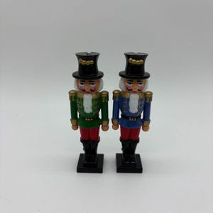 Pode incluir: Dois quebra-nozes decorativos de madeira. Um veste uma jaqueta verde e calças vermelhas, o outro uma jaqueta azul e calças vermelhas. Ambos têm cartolas pretas, cabelo branco e bigodes. Estão em bases pretas.