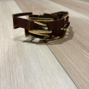 Może przedstawiać: Brązowa skórzana bransoletka z wieloma prostokątnymi klamrami w kolorze złotym. Bransoletka leży na jasnej drewnianej powierzchni. Bransoletka wydaje się nieużywana.