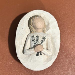 Puede incluir: Figura tallada de una persona con un vestido blanco que sostiene un manojo de hojas verdes. La figura tiene una cabeza de color marrón claro y está colocada sobre una base blanca de forma ovalada. El vestido tiene un patrón texturizado.