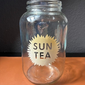 May include: A clear glass jar with a wide mouth, featuring a gold-colored sunburst design with the words "SUN TEA" in the center. The jar is empty and sits on an orange surface, with a dark background.