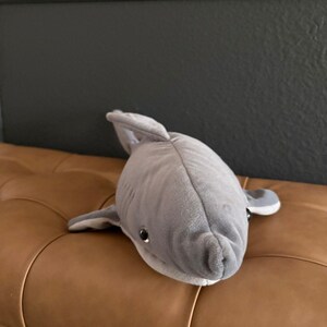 May include: A plush shark toy featuring a soft, light gray exterior with a smooth texture. The stuffed animal has a prominent dorsal fin, side fins, and dark, round plastic eyes. The underside of the shark is a lighter, off-white color.