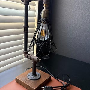 Puede incluir: Lámpara de escritorio de estilo industrial con base de madera y estructura de tubería metálica. La lámpara tiene una jaula de alambre negro alrededor de la bombilla y una bombilla de estilo vintage. La lámpara está enchufada a una toma de corriente.