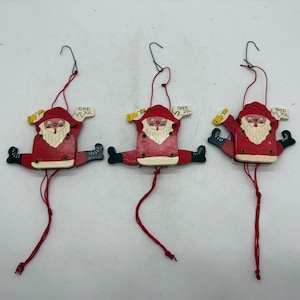 May include: Three hanging wooden ornaments shaped like Santa Claus figures in red suits with white beards. Each figure holds a small sign that reads God Jul and has articulated legs attached by red strings, allowing them to move when pulled.