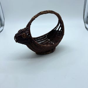 May include: A small, brown woven basket with a handle. The basket has a unique shape, with a rounded base and a handle that arches over the top. The basket is made of tightly woven, dark brown material. The basket is likely used for decorative purposes.