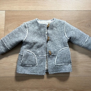May include: A gray long-sleeved jacket featuring a soft white faux-shearling lining, a round neckline, and three wooden toggle closures down the front. The jacket includes two front pockets with exposed white seams along the edges.