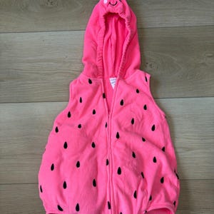 Op de afbeelding: Een roze babykostuum dat op een aardbei lijkt. De outfit met capuchon heeft een rits aan de voorkant en zwarte zaaddetails. De capuchon heeft een groene steel en een lachend gezicht.