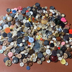 Op de afbeelding: Een grote verzameling knopen in verschillende maten, kleuren en materialen. De knopen omvatten tinten blauw, bruin, zwart, wit en roze. Sommige knopen hebben meerdere gaten, andere een schacht. Er is een verscheidenheid aan vormen en texturen.