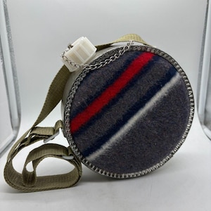 Puede incluir: Una cantimplora verde oliva vintage con una funda de lana con rayas rojas, blancas y azules. La cantimplora tiene un borde de metal, una tapa de plástico blanco y una correa para el hombro de color caqui. Una cadena plateada conecta la tapa a la cantimplora.