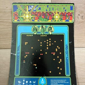 May include: A rectangular Centipede arcade game marquee sign featuring a black background with a green and blue border. The top displays the Centipede logo and colorful mushroom graphics. The center shows game scores and pixelated icons.