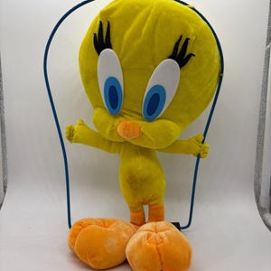 May include: A plush yellow Tweety Bird character with large blue eyes, black eyelashes, and an orange beak. The character is holding a blue wire frame and has two orange plush feet. A classic cartoon character.