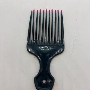 May include: A dark teal Afro pick with a handle and a row of tines. The tips of the tines are painted magenta. The pick is designed for styling and detangling textured hair.