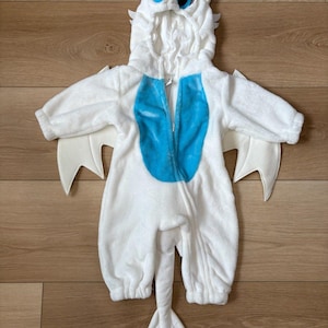 May include: White plush baby costume with a dragon design. The hooded jumpsuit features a blue chest panel, attached wings, and a tail. The hood has large, blue-eyed dragon face details.
