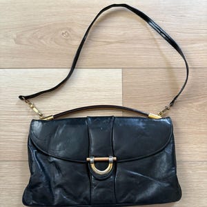 以下が含まれることがあります： 黒い革のハンドバッグ。長いショルダーストラップが付いています。バッグは、金色の金属と木材のアクセントが付いたフラップクロージャーが特徴です。バッグは長方形で、上部にハンドルが付いています。