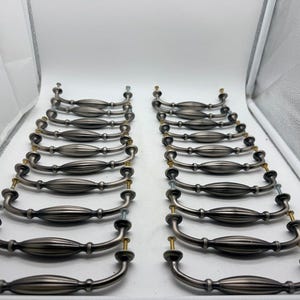 May include: A collection of sixteen matching metal drawer pulls arranged in two parallel rows. Each pull features a curved, ribbed center section with a brushed silver finish and includes two small gold-toned mounting screws at the ends.