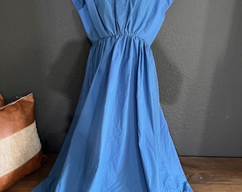 May include: A light blue dress with short sleeves and a gathered waist. The dress is displayed on a white mannequin and has a flowing skirt. The background is a dark gray wall and a wooden bench.