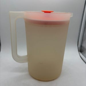 Può includere: Una brocca vintage in plastica smerigliata con manico e coperchio. La brocca è di colore beige chiaro, con un coperchio arancione e una piccola linguetta arancione. Il manico è trasparente. Ideale per servire bevande.