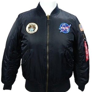 May include: Black bomber jacket with a gold zip, featuring NASA and Space Command patches. Includes a red "Remove Before Flight" tag and a small American flag patch. A classic outerwear piece.