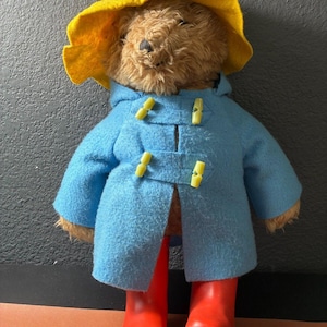 Puede incluir: Un oso de peluche con un sombrero amarillo, un abrigo azul claro con broches amarillos y botas de agua rojas. El oso es un juguete infantil clásico.