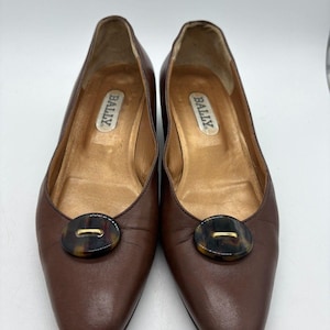 Könnte beinhalten: Ein Paar braune Bally-Flats aus Leder. Jeder Schuh hat einen Knopf im Schildpatt-Stil mit goldenem Akzent. Das Bally-Logo ist in jedem Schuh sichtbar. Die Schuhe werden auf einer weißen Oberfläche präsentiert.