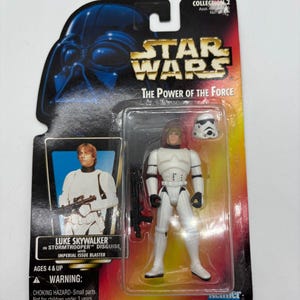 Op de afbeelding: Een Star Wars actiefiguur van Luke Skywalker in Stormtrooper vermomming, verpakt op een kaart. De figuur is wit met zwarte accenten en wordt geleverd met een blaster en een Stormtrooper helm. De verpakking toont het Star Wars logo en de tekst "The Power of the Force."