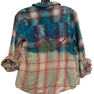 May include: A plaid button-down shirt in shades of red, white, and blue, with a turquoise-dyed back panel. The back features the word "Hello" in pink glitter script above a green glitter leaf design. The sleeves are rolled up.