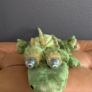 May include: A green plush alligator toy featuring a textured back with raised ridges and two prominent, protruding eyes. The eyes are encased in clear, spherical plastic domes with detailed, realistic irises and pupils, set within soft fabric sockets.